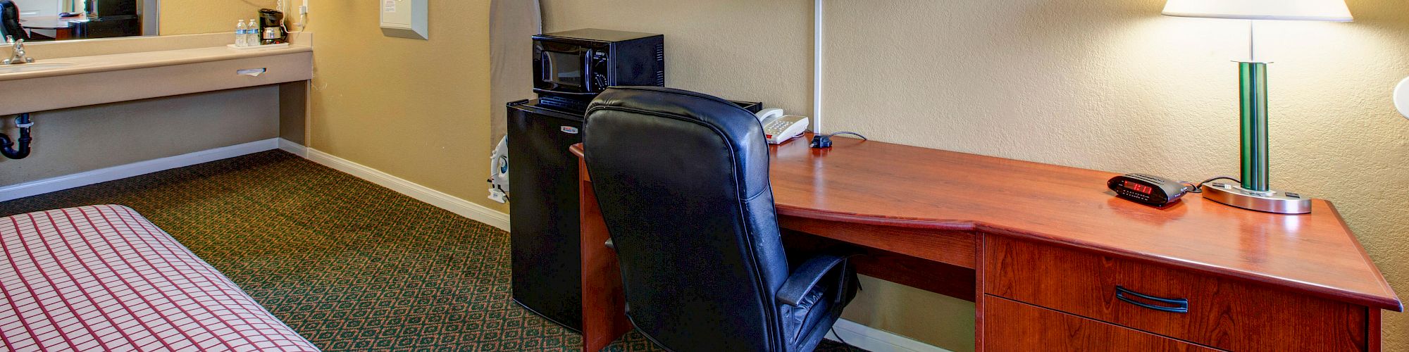 A hotel room with a bed, TV, desk, chair, microwave, lamp, and a vanity area with a sink and mirror.