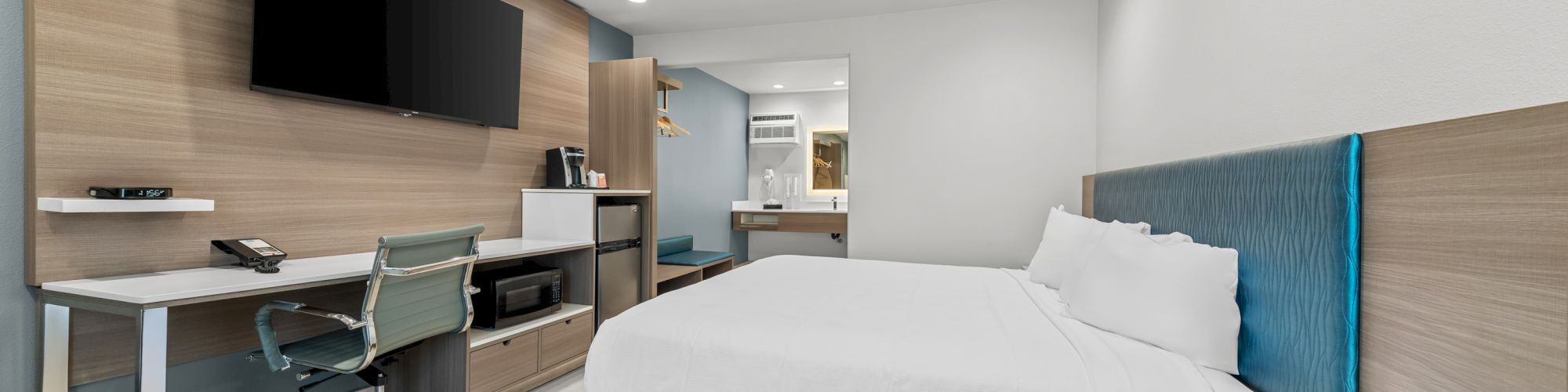 A modern hotel room with a large bed, blue headboard, desk with chair, and wall-mounted TV.