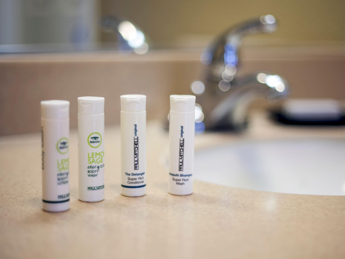 The image shows four white cylindrical toiletry bottles on a beige countertop near a sink with a shiny faucet in the background.