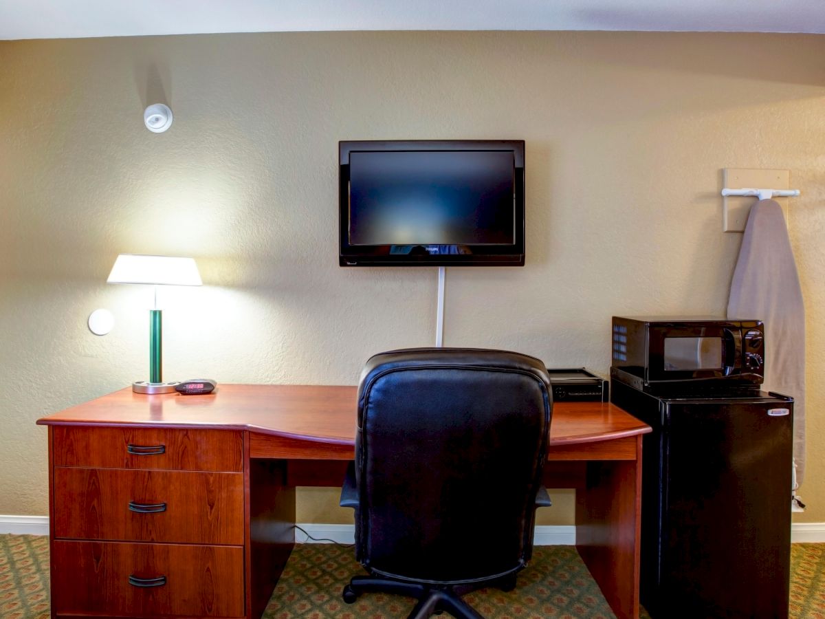 The image shows a small workspace with a wooden desk, office chair, wall-mounted TV, a desk lamp, mini refrigerator, microwave, and coat rack on the wall.