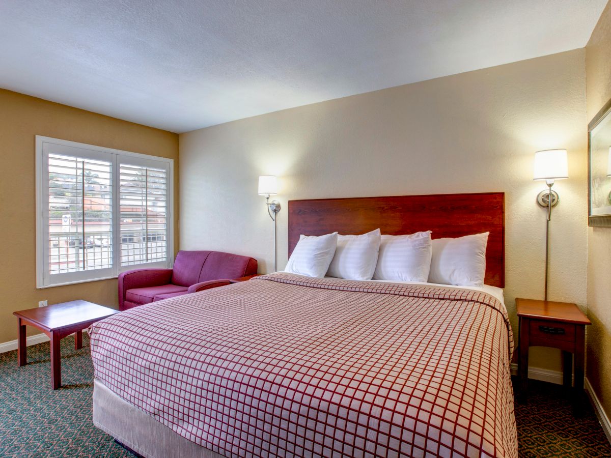 A well-lit hotel room with a large bed, two bedside tables, a reading chair, coffee table, window with blinds, and simple decor.