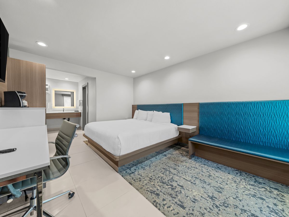 A modern hotel room with a bed, blue tufted bench wall, desk, chair, and bright ceiling lights.