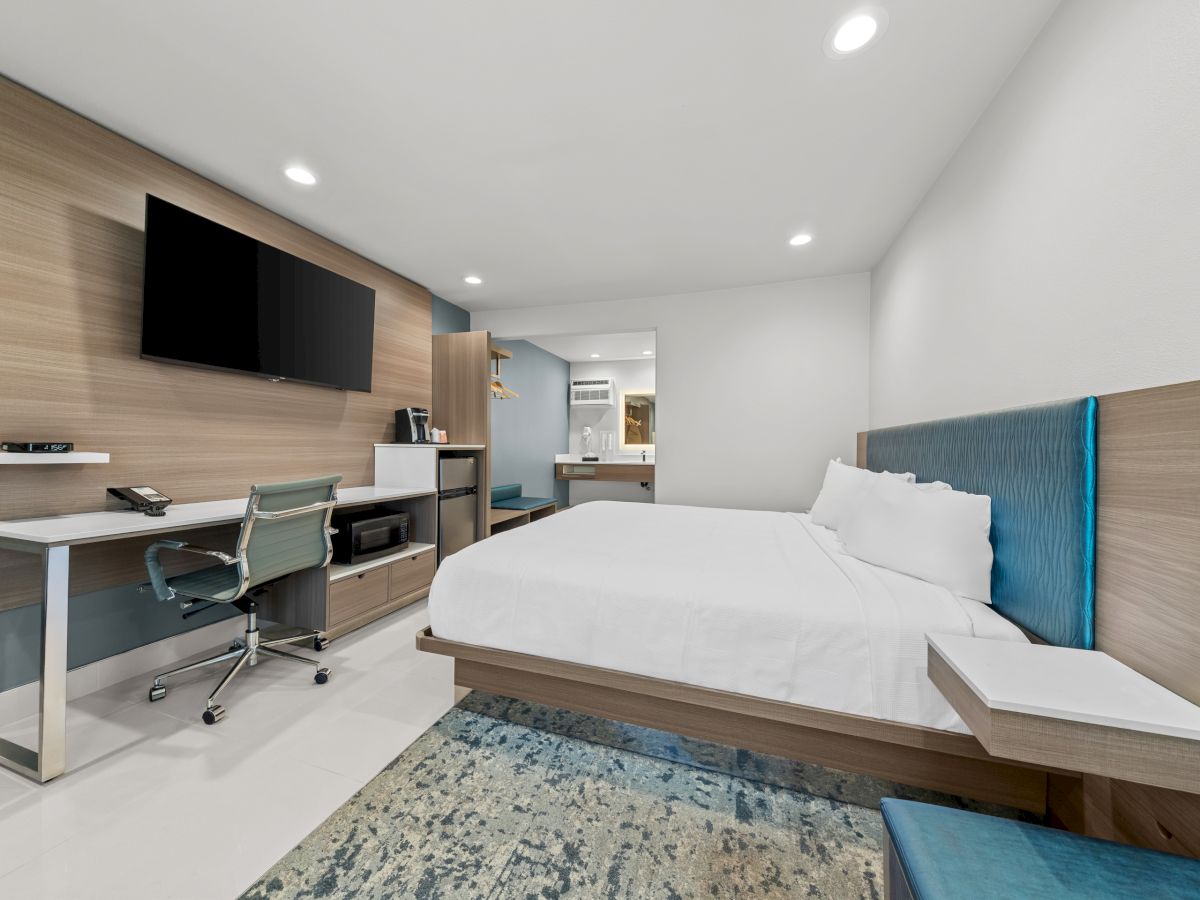 A modern hotel room with a bed, desk, chair, wall TV, and a blue upholstered headboard.
