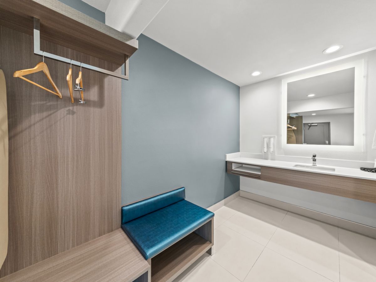 Modern bathroom with a vanity, wall mirror, towel on a rack, hanging clothes, and a blue bench for getting ready.