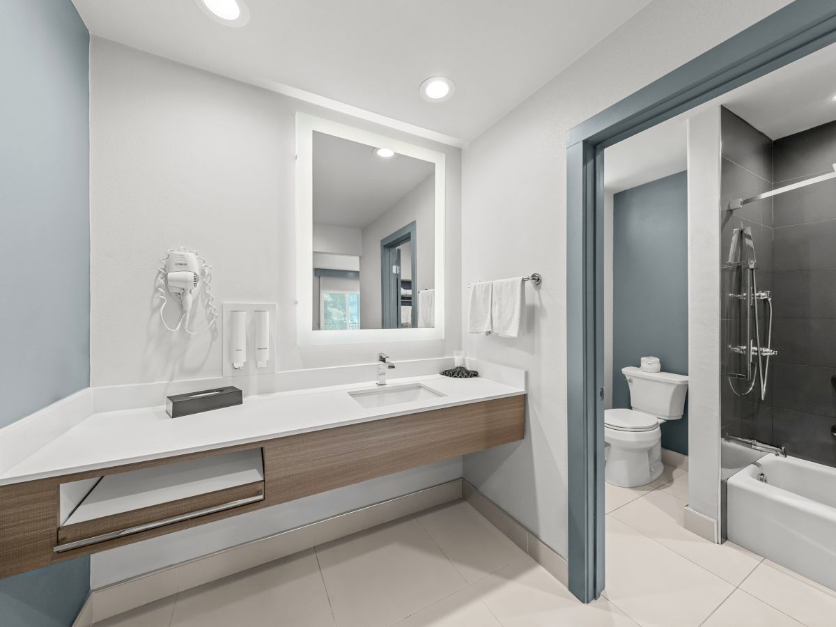 A modern bathroom with a vanity sink, mirror, towels, toilet, and a glass shower/tub area under bright ceiling lights.