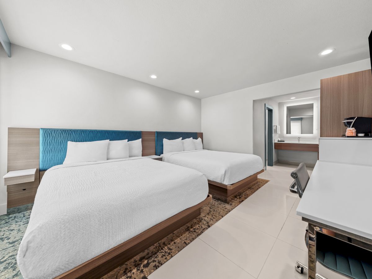 Bright modern hotel room with two twin beds, blue headboards, tiled floor, and a kitchenette with coffee maker.