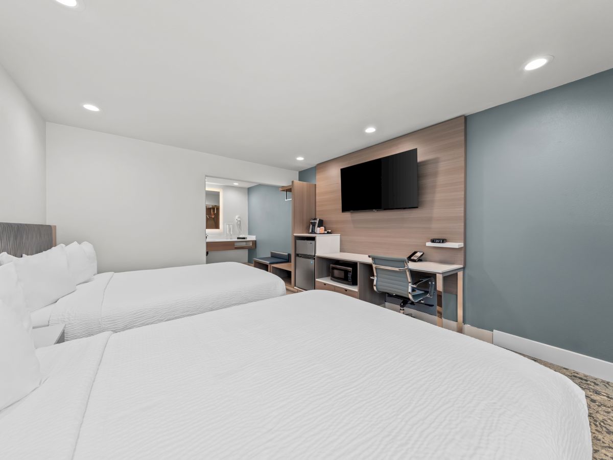 A modern hotel room with a large white bed, desk and chair, flat-screen TV, and a compact kitchenette area in the back.