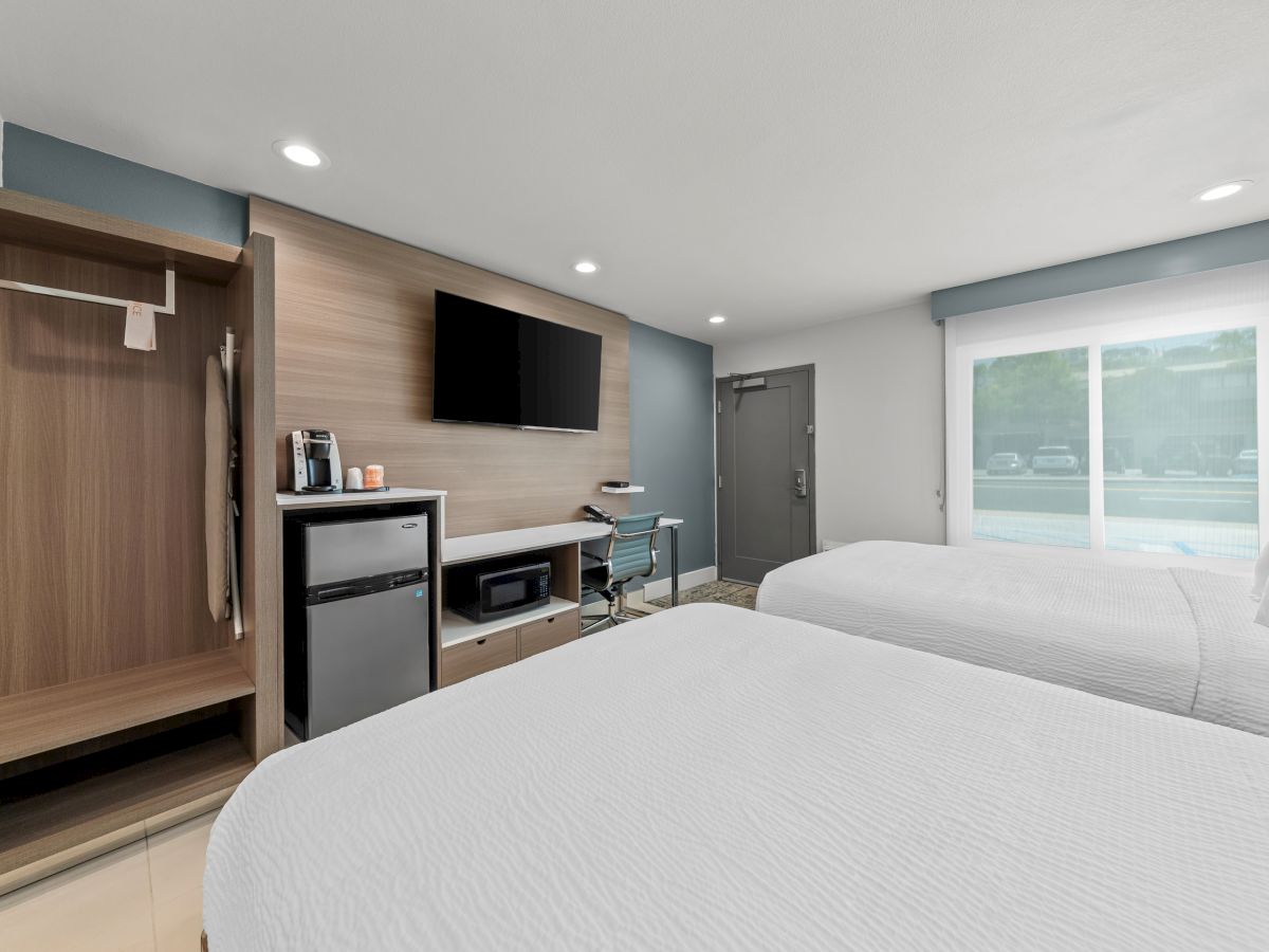 Modern hotel room with two beds, flat-screen TV, mini-fridge and microwave, desk area, and a large window with blinds.