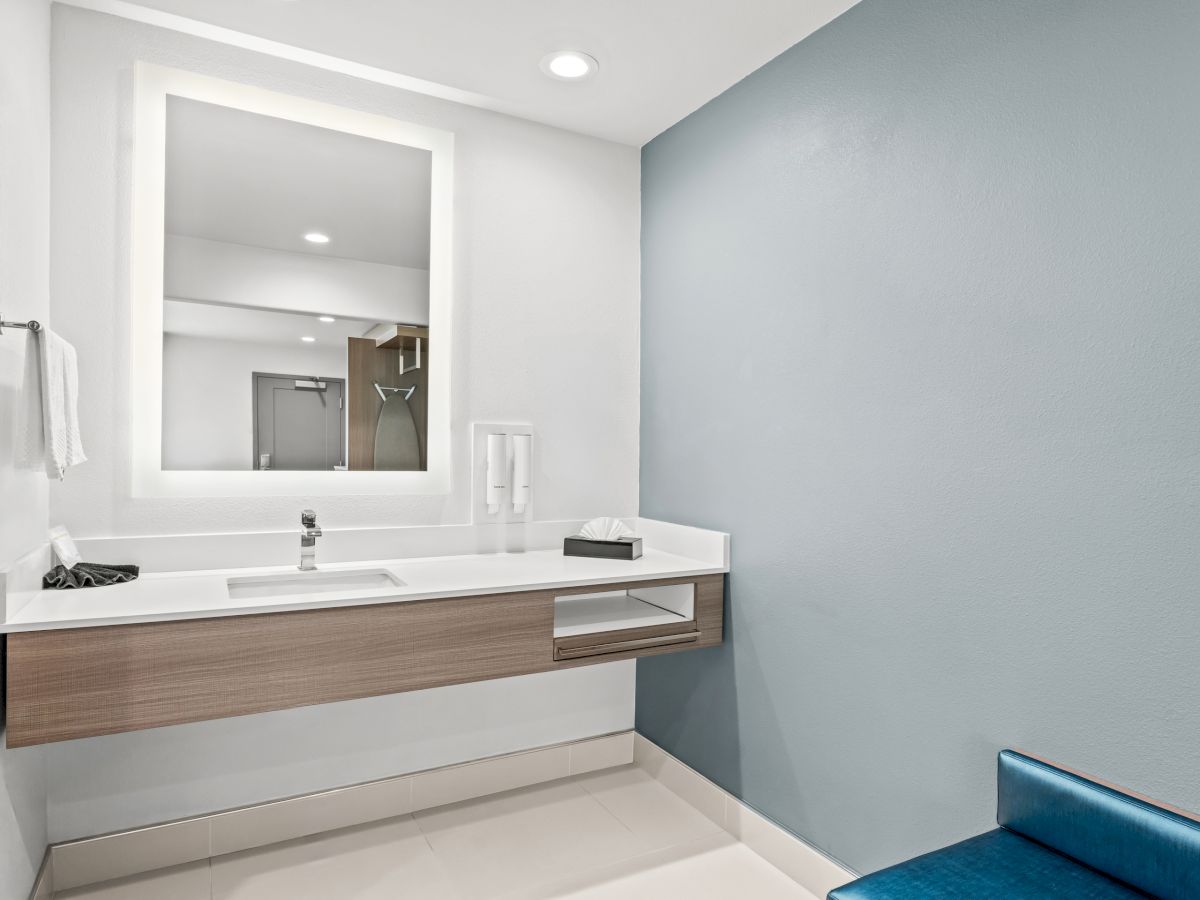 A modern bathroom with a wall-mounted vanity, white sink, towel, dispenser bottles, and a flat mirror under recessed lights.