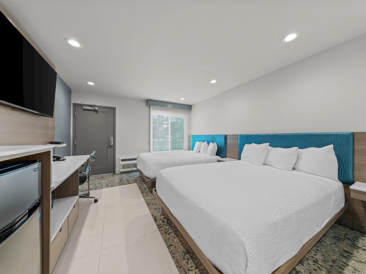 Modern hotel suite with two white beds, blue headboards, a TV, desk setup, and large window under ceiling lights.