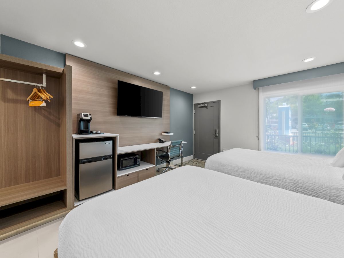 A modern hotel room with two white beds, a desk and chair, a wall TV, and a large window with blinds, plus a coffee station.