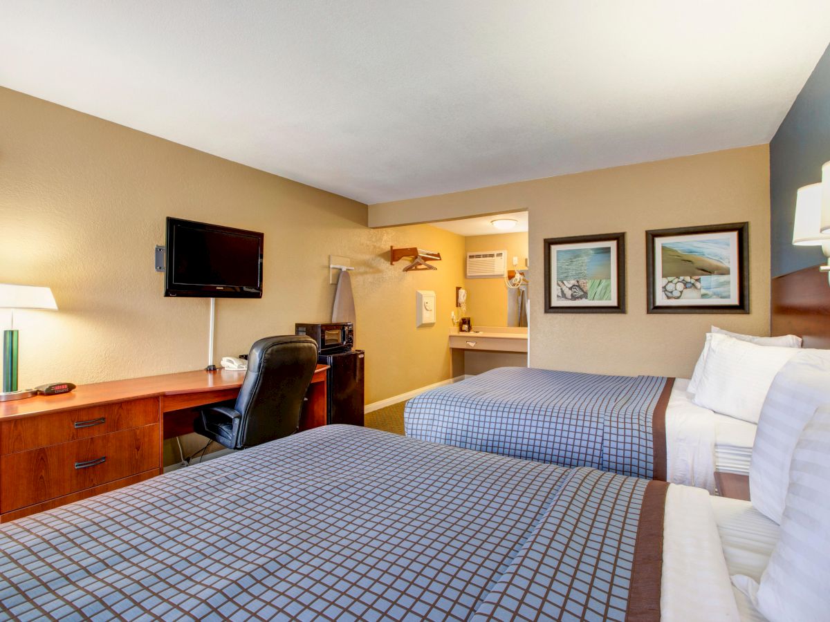 A hotel room with two beds, a desk, chair, TV, wall art, and a lamp; open doorway leading to a bathroom area.