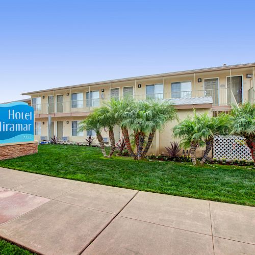 A two-story building identified as "Hotel Miramar" with a sign on a grassy lawn, small palm trees, and a clear sky in the background.