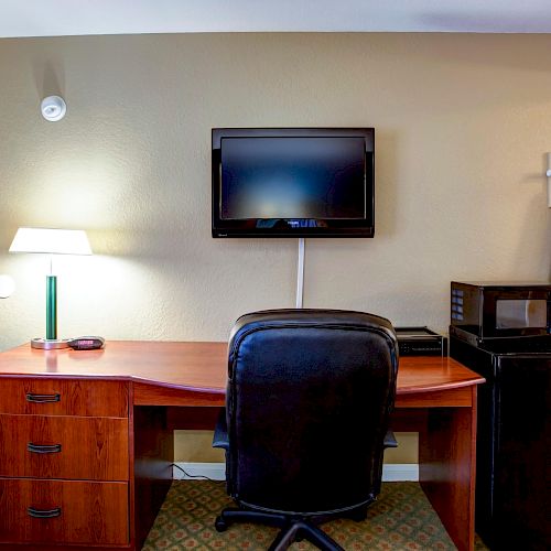 A desk with a lamp and phone, a chair, a wall-mounted TV, hanging clothes, a microwave, and a mini-fridge in a room.