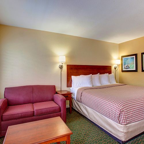 The image shows a hotel room with a king-sized bed, a red sofa, a wooden coffee table, two framed pictures, and bedside lamps.