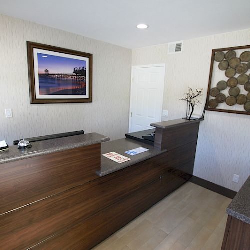 A reception area with a wooden desk, two wall pictures, a sign, some papers, and decorative items atop a countertop ending the sentence.