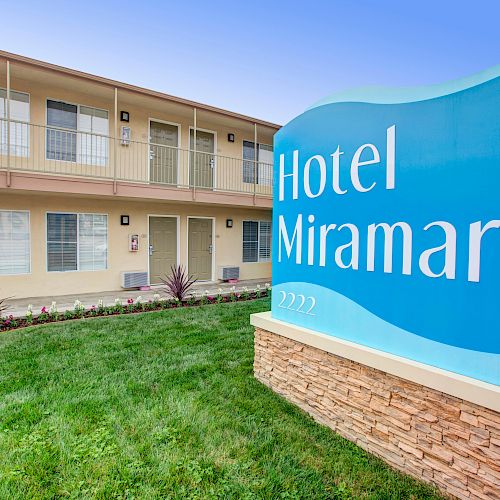 The image shows the exterior of a two-story hotel with a blue sign that reads "Hotel Miramar" on a neatly landscaped lawn.