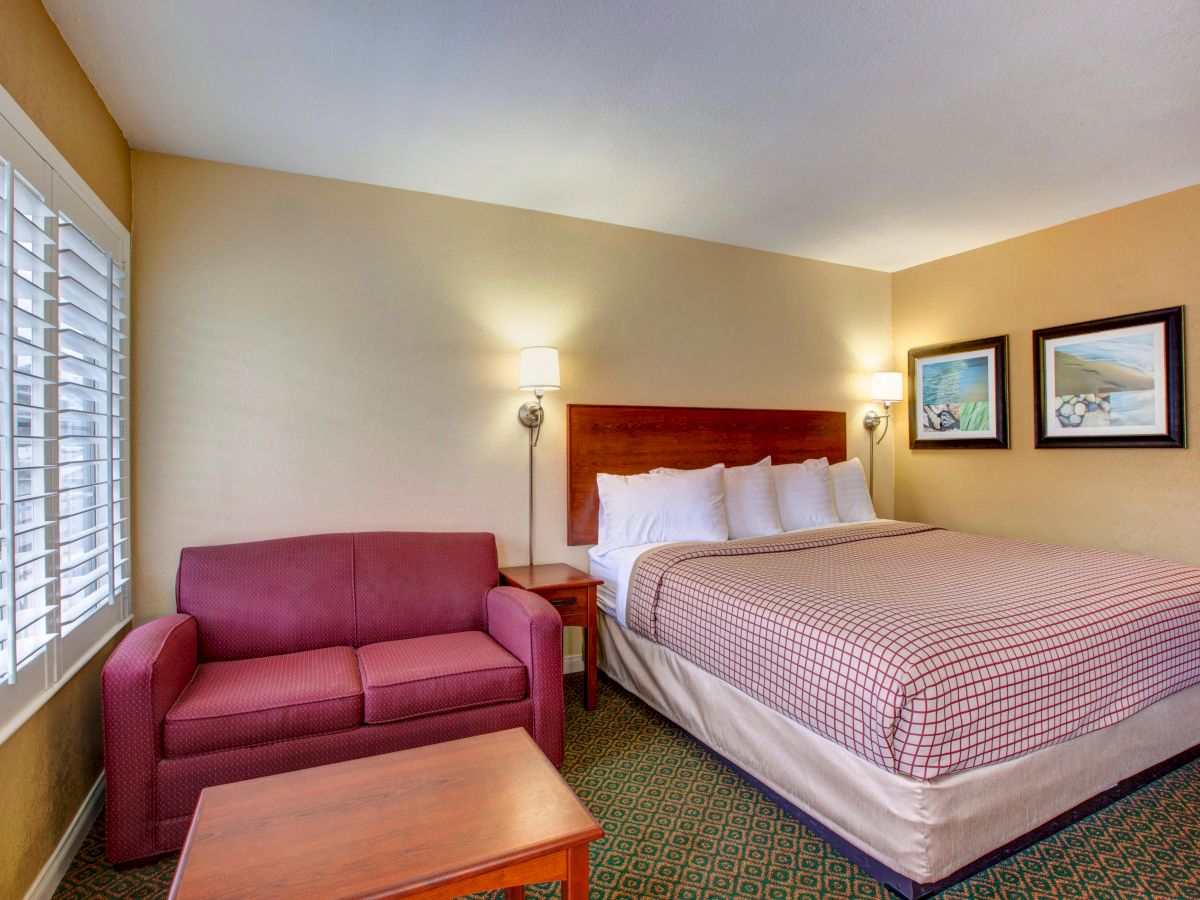 A hotel room features a bed with checkered bedding, a red sofa, wooden tables, wall art, and lamps, with natural light streaming in through shutters.