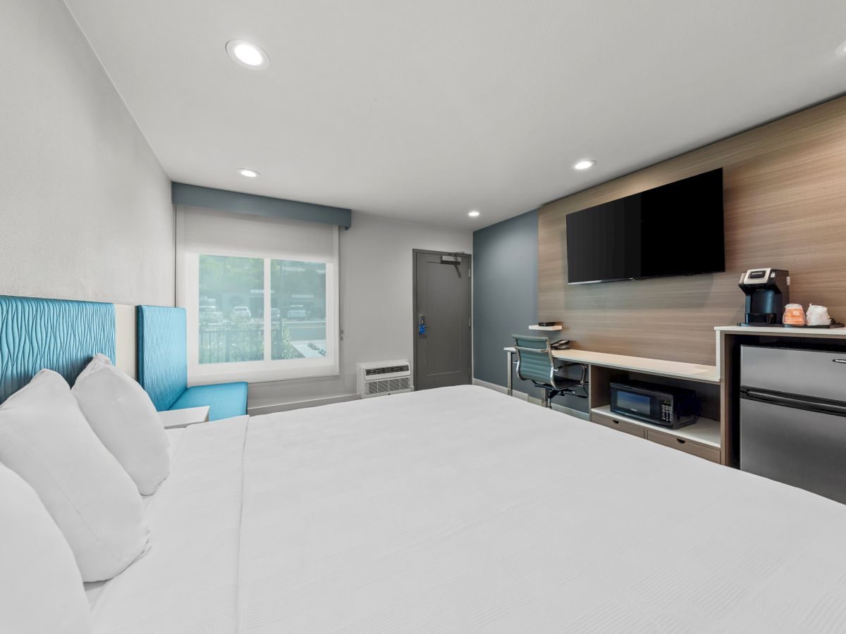 A modern hotel room with a large bed, window, desk with TV, coffee maker, and mini-fridge.