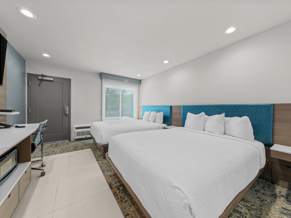 A modern hotel room with two beds, a kitchenette counter, and a gray door under ceiling lights.