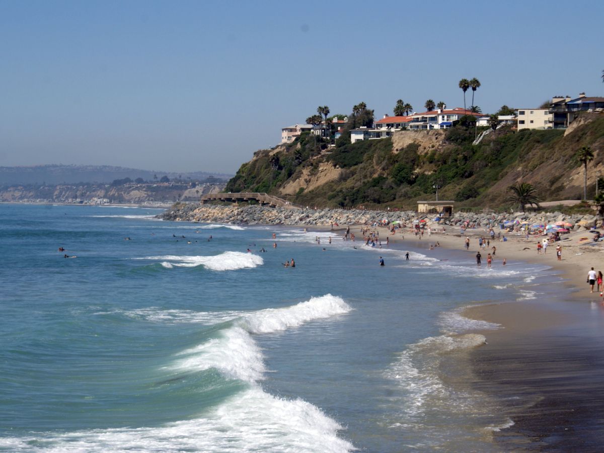 A scenic beach with waves crashing on the shore, people enjoying the sun and water, and houses on a hillside in the background, ending the sentence.