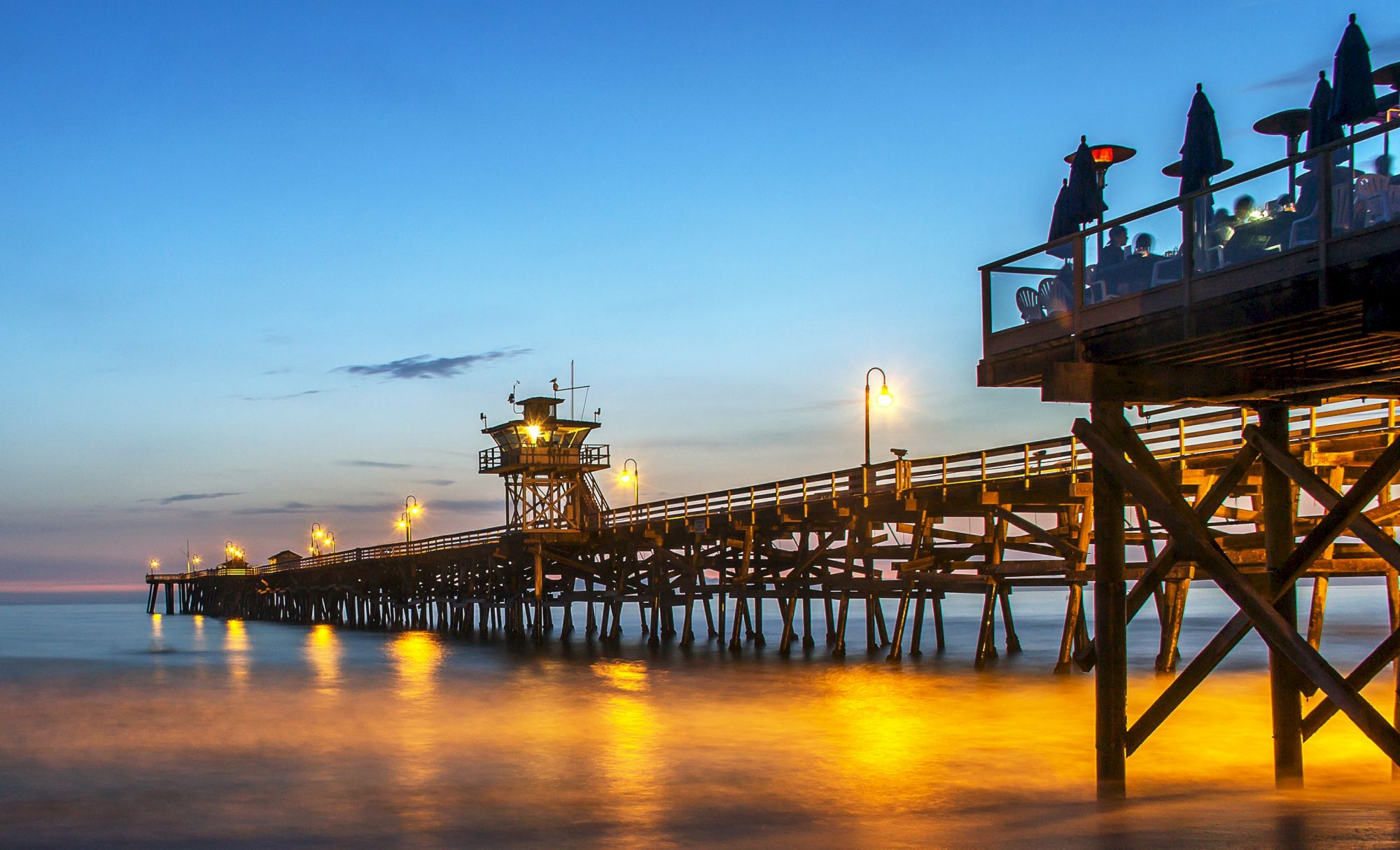 A wooden pier extends over calm water at sunset, with lights illuminating its length. There's a seating area with umbrellas on the right side of the image.