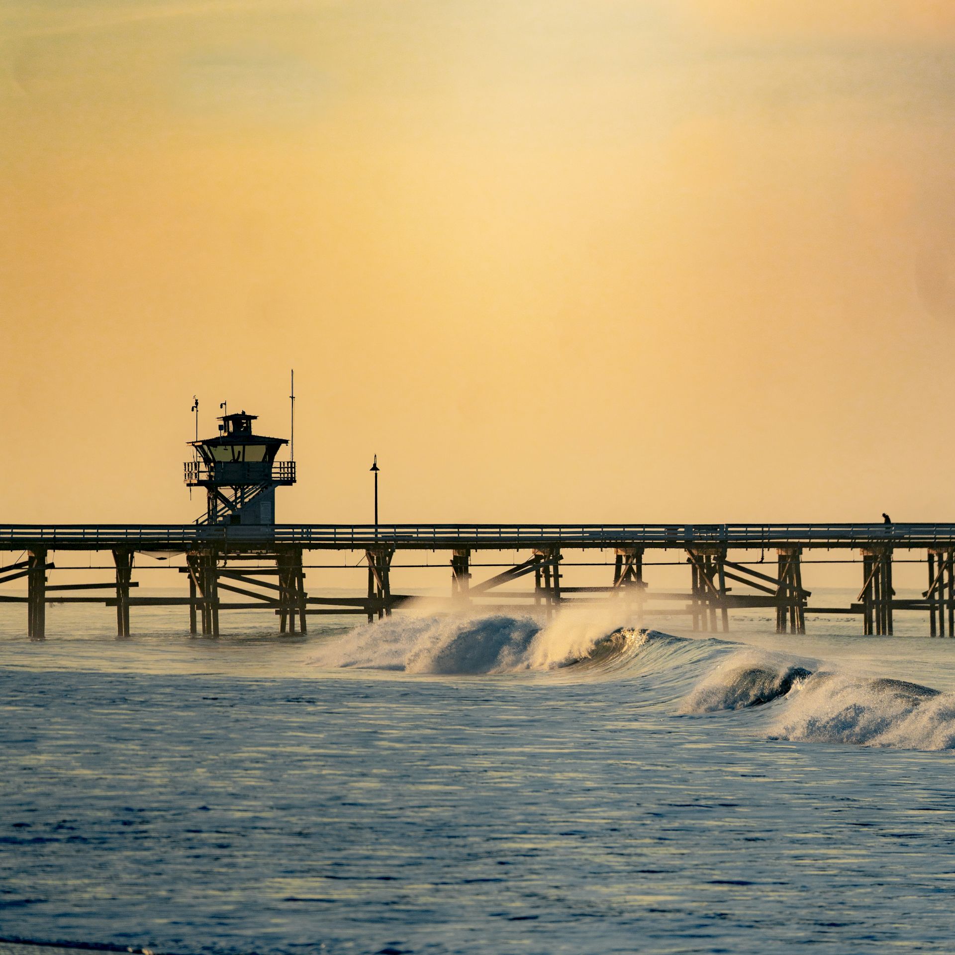 A wooden pier extends over the ocean with small waves in the foreground and a soft sunset in the background.