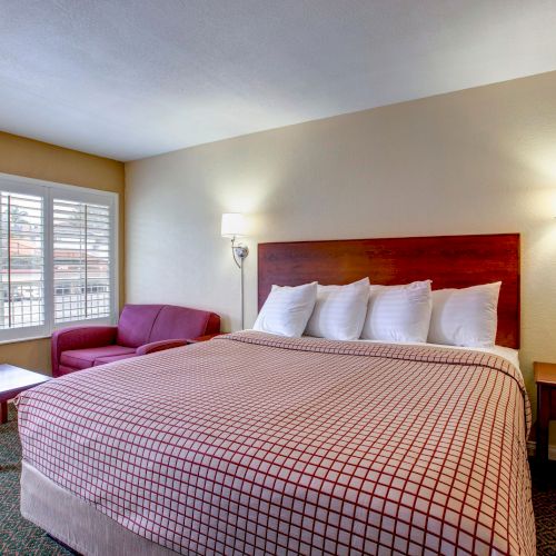 A cozy hotel room with a king-sized bed, decorative pillows, two lamps, a red sofa, a coffee table, and a window with shutters.