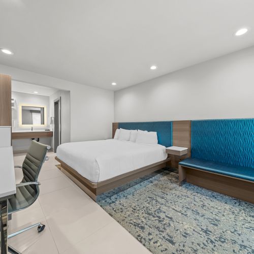 Modern hotel room with a white bed, blue upholstered bench, desk area, and recessed ceiling lights.