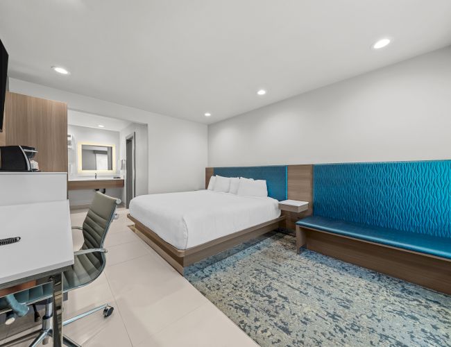 Modern hotel room with a white bed, blue upholstered bench, desk area, and recessed ceiling lights.