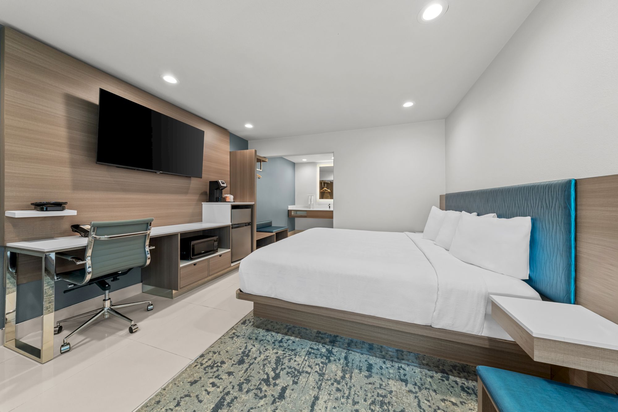 A modern hotel room with a large bed, desk chair, flat-screen TV, and bright ceiling lights.