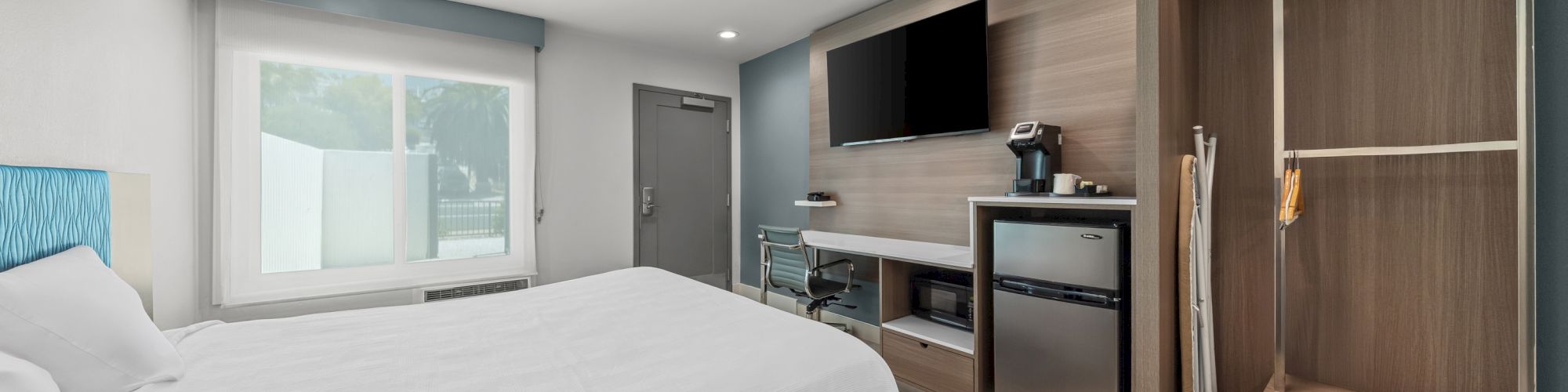 A modern hotel-style room with a large bed, window, TV wall, minibar area, and built-in shelves and storage.
