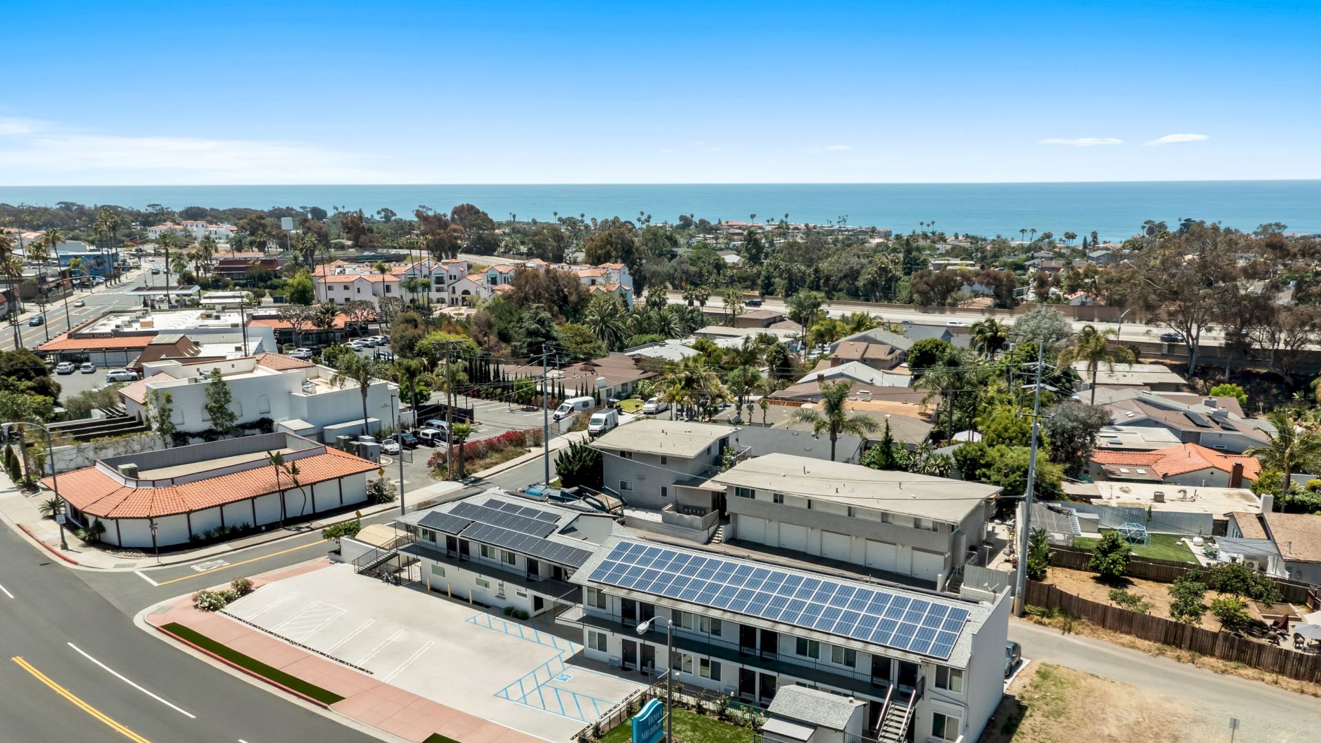 Aerial view of a coastal neighborhood with a parking lot and buildings, including rooftops covered in solar panels, under a clear sky.
