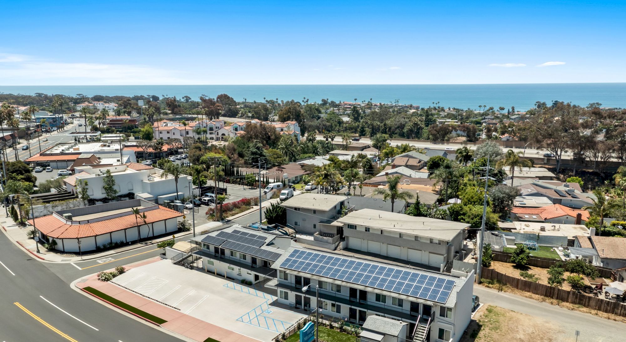 Aerial view of a coastal neighborhood with a parking lot and buildings, including rooftops covered in solar panels, under a clear sky.