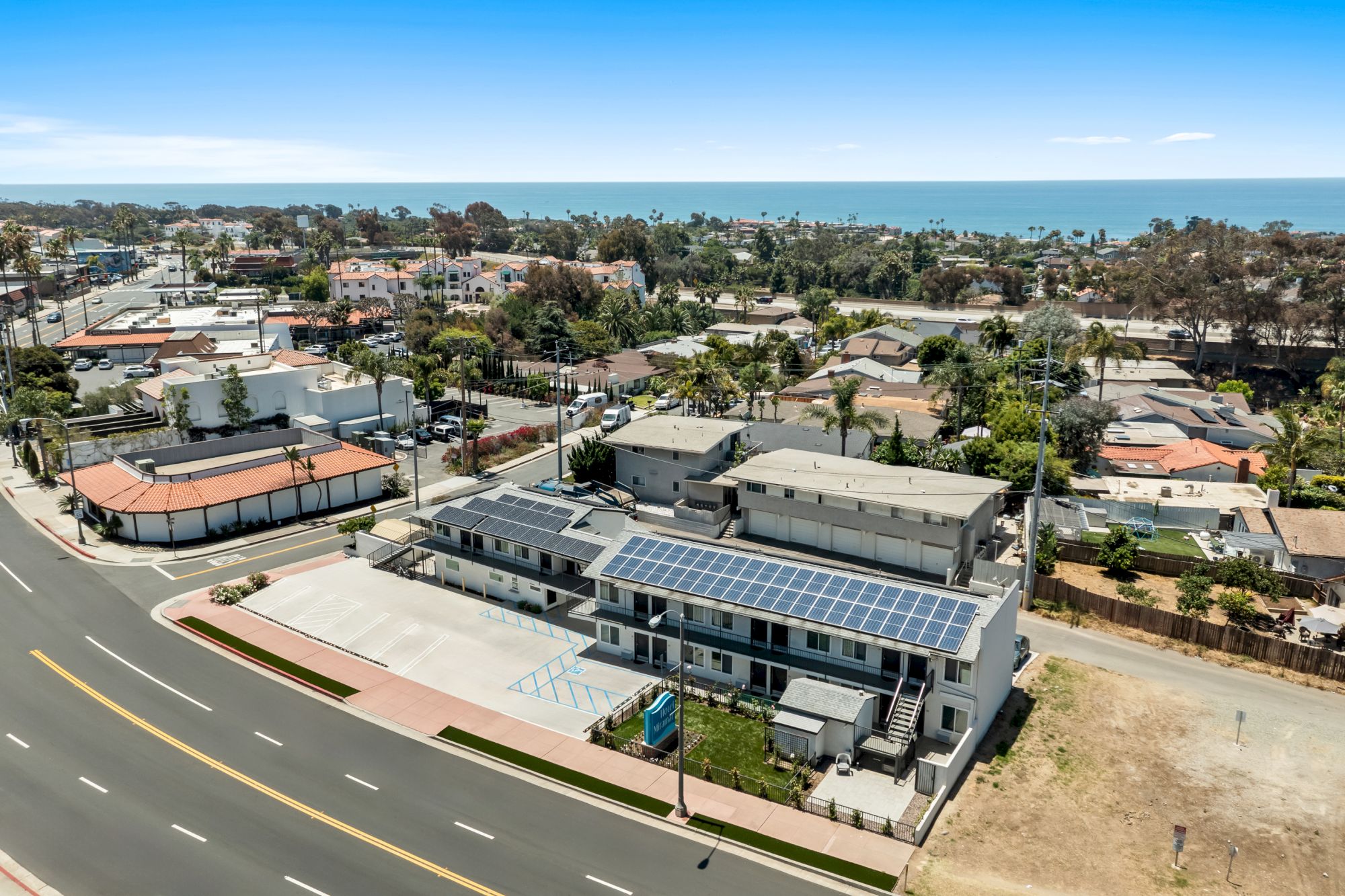 Aerial view of a coastal neighborhood with a parking lot and buildings, including rooftops covered in solar panels, under a clear sky.
