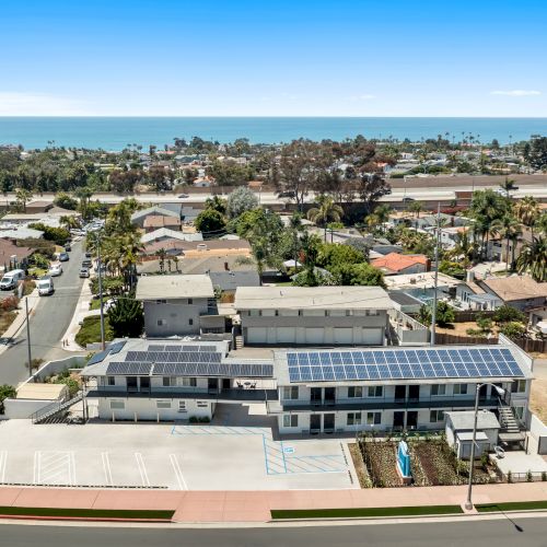 An aerial view of a coastal neighborhood with a large building rooftop solar panels, palm trees, roads, and the blue ocean horizon.