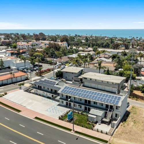 Aerial view of a coastal town with roads and homes, plus a building rooftop covered in solar panels under a clear blue sky.