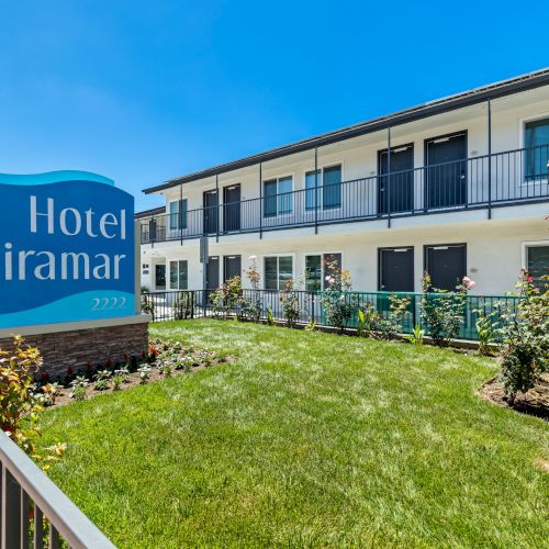 A sunny exterior of the Hotel Miramar, with a blue sign and landscaped lawn beside a multi-story building.