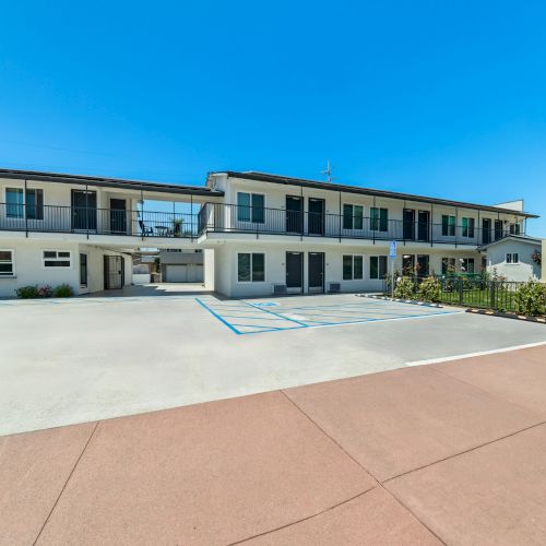 Exterior of Hotel Miramar with a courtyard, parking area lines, and motel-style rooms under a bright blue sky.