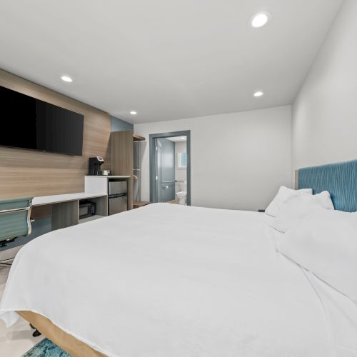 Modern hotel room with a large bed, desk and chair, flat-screen TV, kitchenette counter, and an open bathroom doorway.