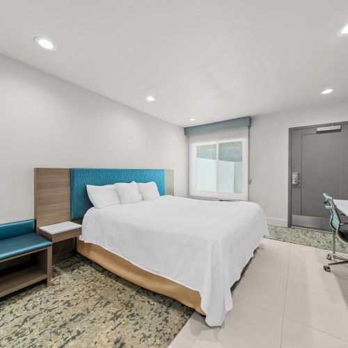 A modern hotel bedroom with a king bed, blue headboard, window, gray door, desk, and built-in TV.