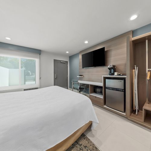 Modern hotel room with a large bed, window, wall TV, mini-fridge/coffee station, and built-in cabinets beside the bathroom.