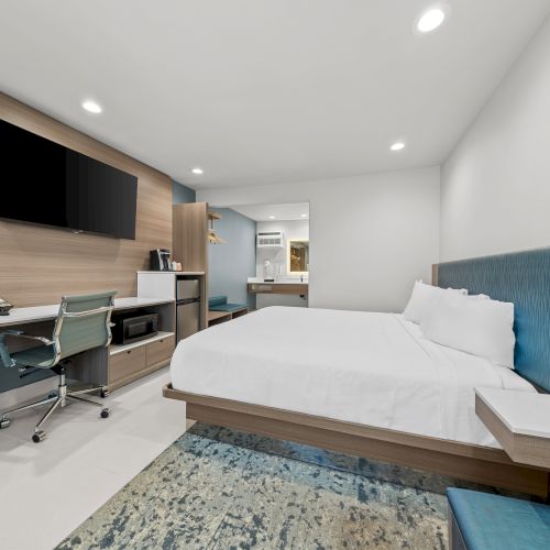 A modern hotel room with a large bed, TV on the wall, a desk with chair, and bright recessed lighting.