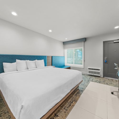 A modern hotel room with a large bed, desk, chair, window, and a gray door.