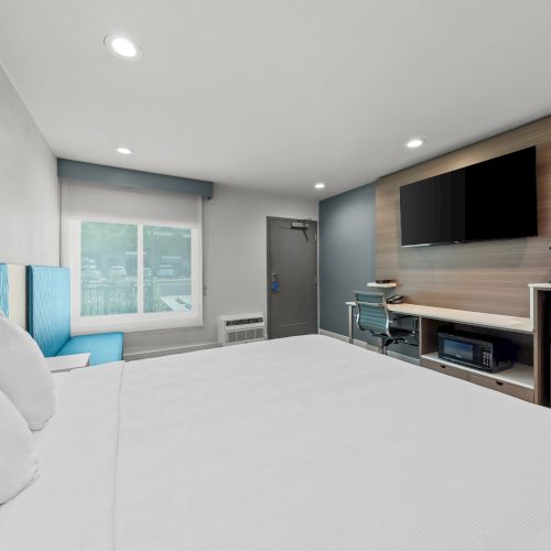 A modern hotel room with a large bed, big window, desk with chair, wall-mounted TV, and a coffee station.