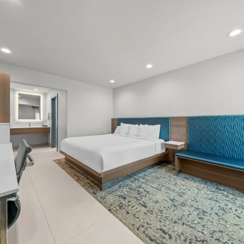 A modern hotel room with a neatly made bed, blue upholstered headboard, tiled floor, and a small kitchenette area.