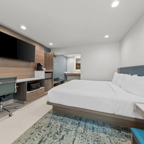 A modern hotel room with a large bed, desk and chair, flat-screen TV, built-in storage, and recessed ceiling lights.