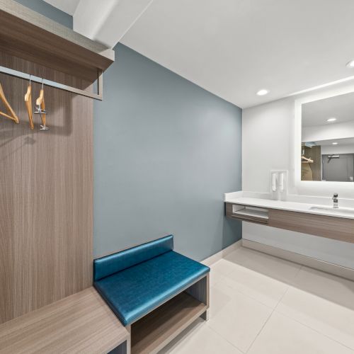 A modern dressing room with coat hangers, a blue bench, and a vanity sink mirrored wall.
