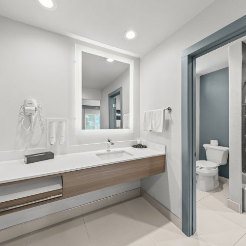 A modern bathroom with a vanity sink, mirror, toilet, and a glass shower/bath with showerhead and towels hanging.
