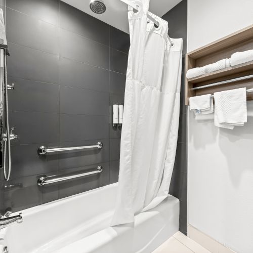 A modern bathroom with a glass shower, tub, white shower curtain, chrome fixtures, and neatly folded towels on a wooden shelf.